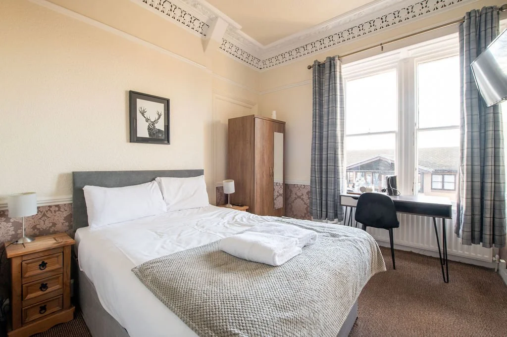 A bedroom with a bed, nightstands, a wardrobe, a desk, a chair, and a window with curtains, with sunlight coming in.