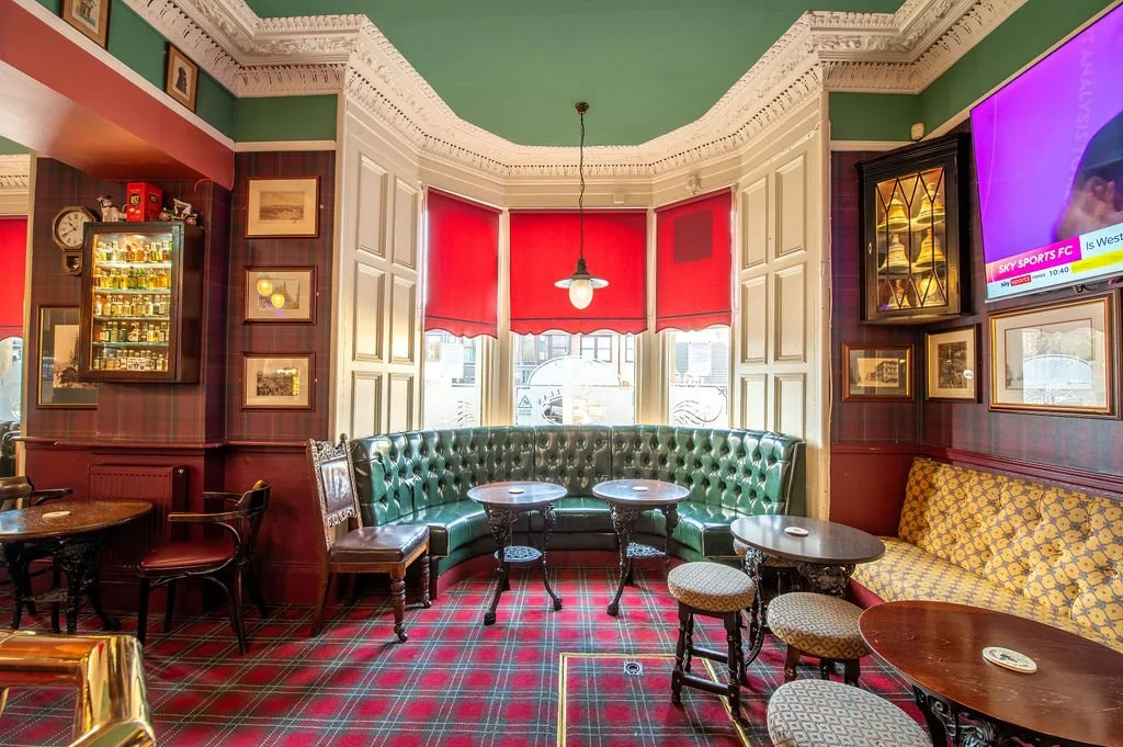 Interior of a cozy pub or restaurant with green leather banquette seating, red window shades, wooden tables, patterned stools, plaid carpet, and framed pictures on wood-paneled walls.