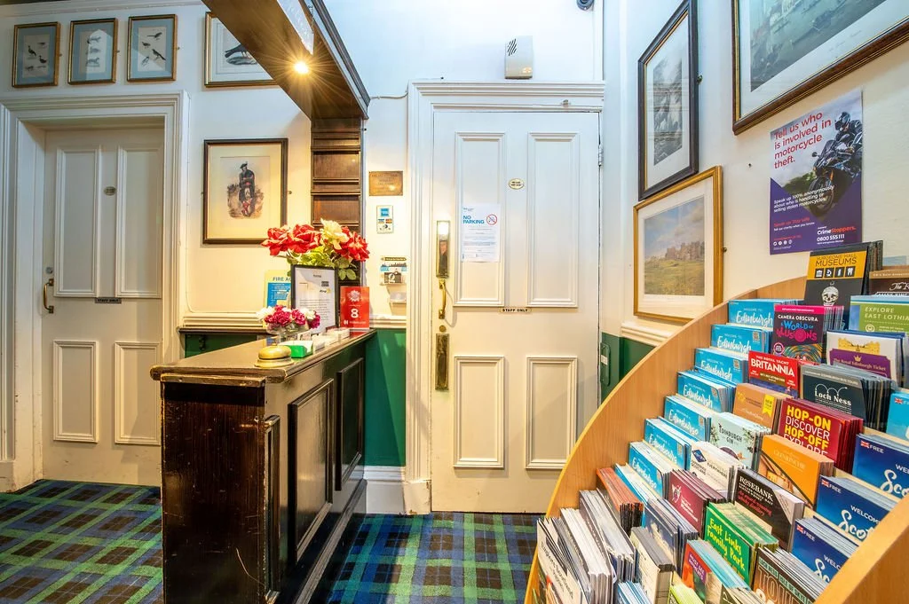 Front desk area of a visitor center with a brochure stand on the right, framed pictures on the walls, a door, and a small counter decorated with flowers.