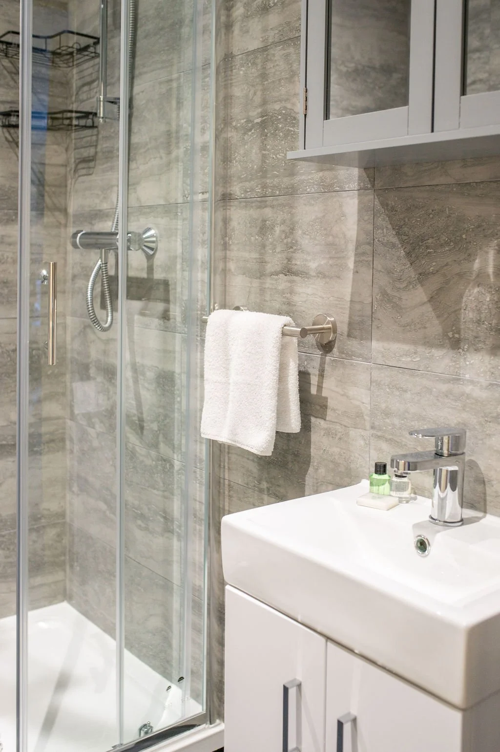 Modern bathroom with glass shower enclosure, white towel on the towel bar, small vanity with soap, lotion, and toothbrush holder, and gray tiled walls.