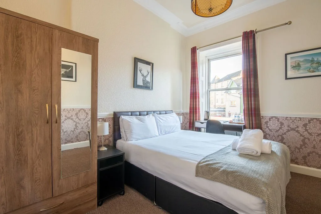 A cozy bedroom with a bed, nightstand, wardrobe, desk, and chair, illuminated by daylight through a window with red checkered curtains.