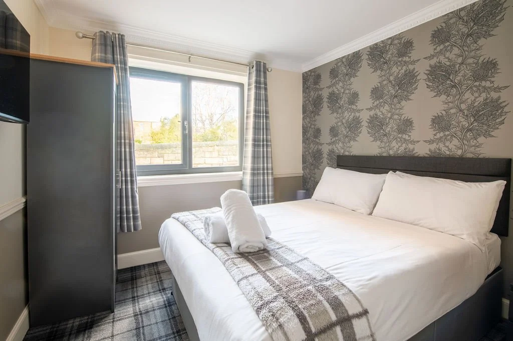 A well-made bed with white linens and pillows, featuring two rolled towels on a plaid blanket at the foot, in a bedroom with a window, plaid curtains, a dark headboard, and patterned wallpaper.