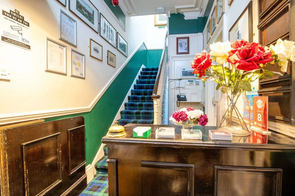 Interior of a reception or lobby area with a wooden counter, large vase with red and white artificial flowers, framed pictures on the wall, a staircase with a plaid carpet, and a wall-mounted telephone.