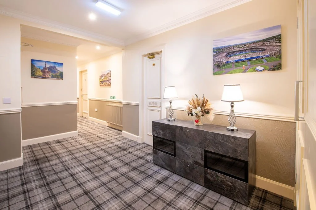 Hallway with framed landscape photos, a marble console table with two lamps and a floral arrangement, and plaid carpet.