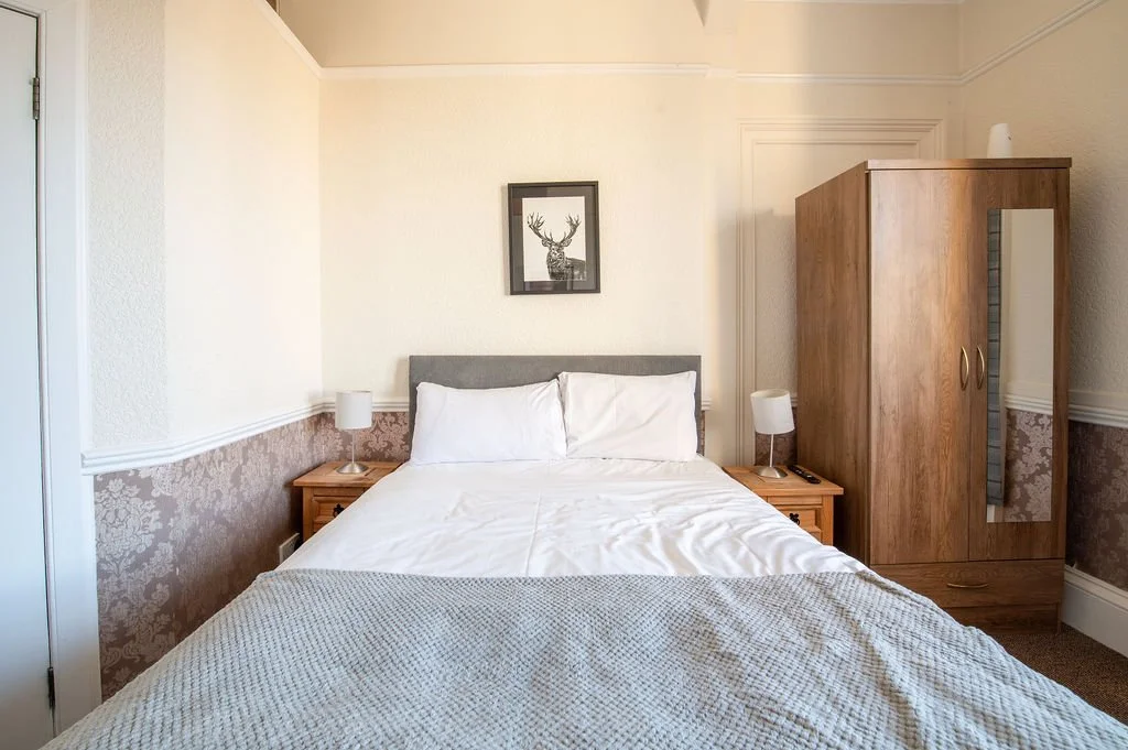 A neatly made bed with white sheets and pillows, two bedside tables with lamps, a framed picture of a deer above the headboard, a wooden wardrobe with a mirror, and a beige wall with decorative wallpaper on the lower half.