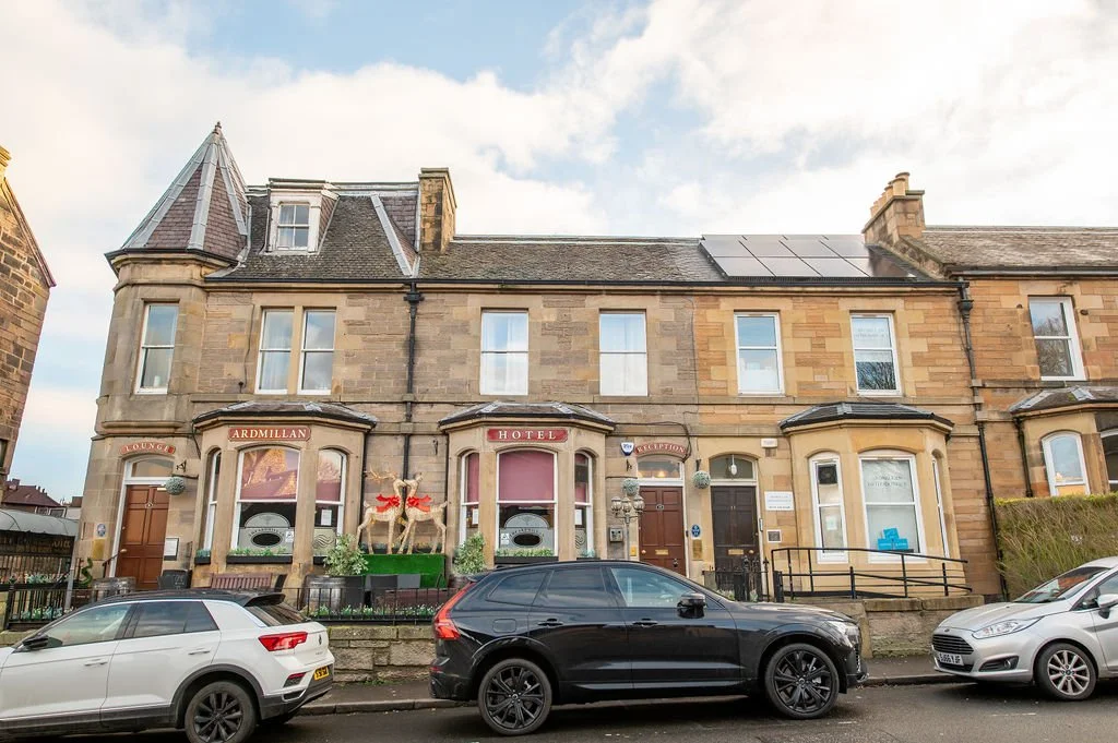 A row of historic stone buildings with a hotel sign, decorated with reindeer holiday figures and greenery, parked cars in front, and a cloudy sky above.