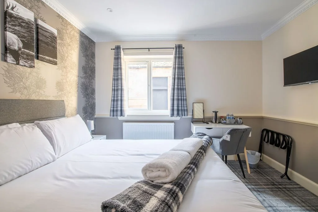 A cozy hotel room with a double bed, a small side table, a window with blue checkered curtains, a desk with a chair, a mirror, a tray with water bottles, and a wall-mounted flat-screen TV.