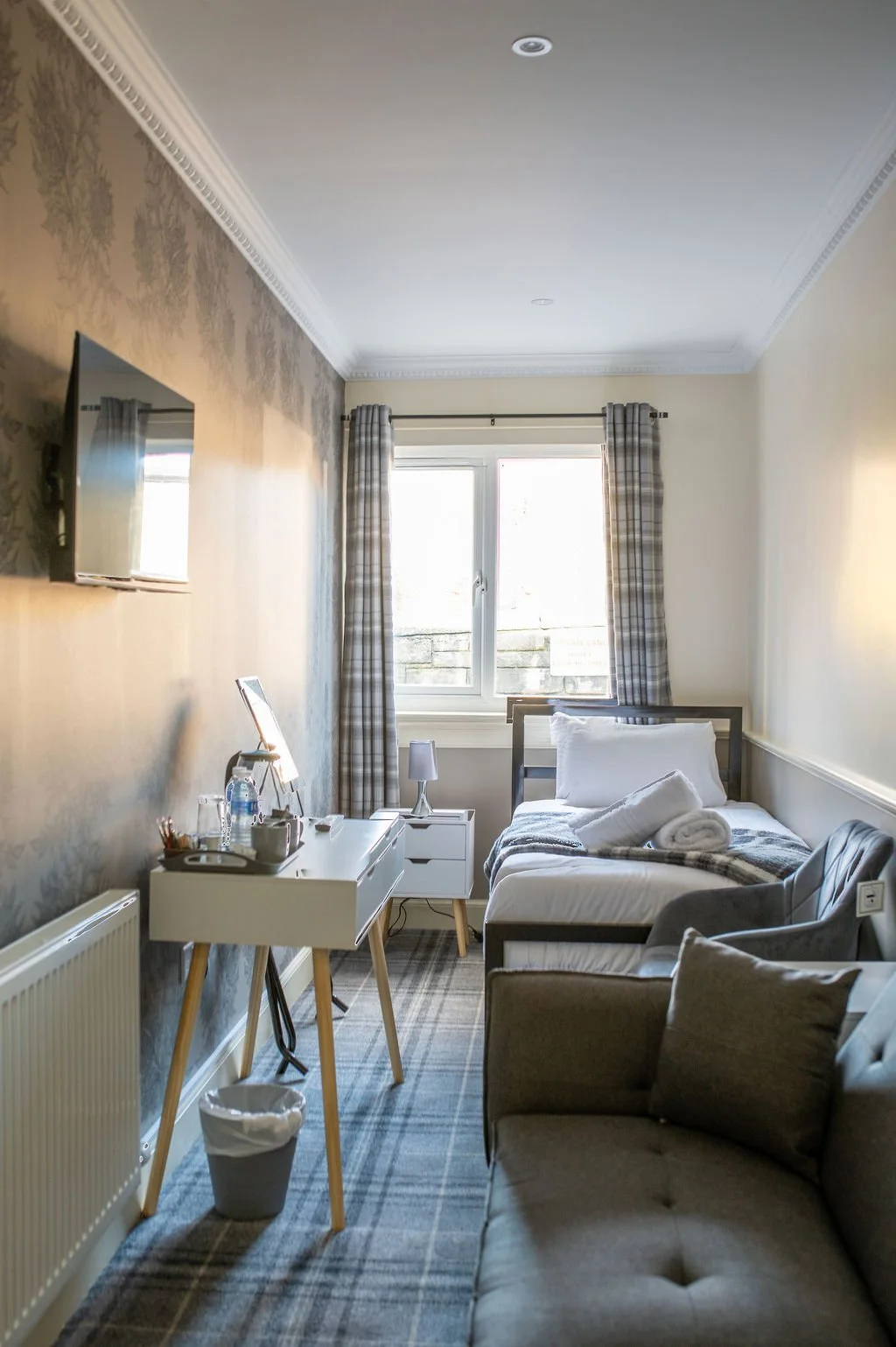 A compact hotel room featuring a single bed with pillows and a blanket, a small gray sofa, a white desk with a tray and water bottles, a window with plaid curtains, a wall-mounted TV, and a radiator.