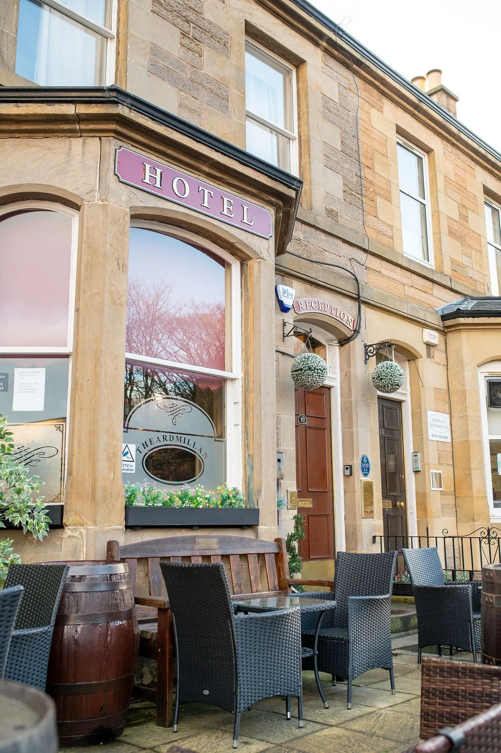 Exterior of a hotel with outdoor seating, wooden barrels, black chairs, and a window with flower boxes.