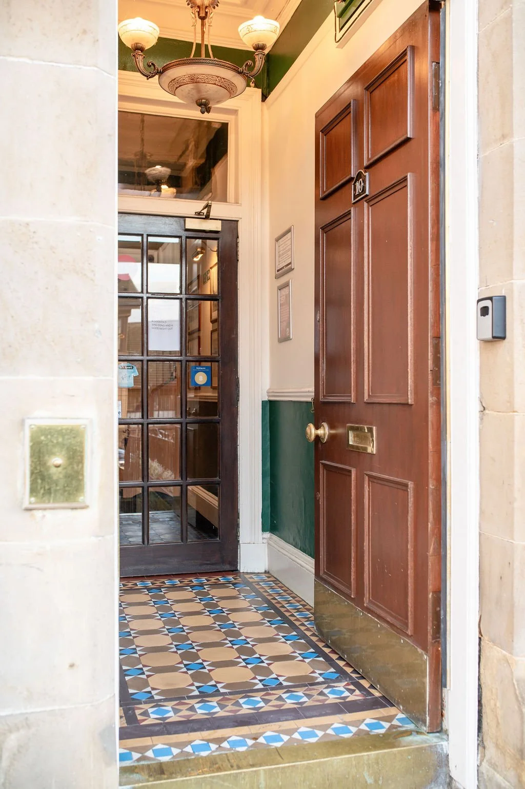 The image shows a view of a building entrance with an open wooden door and a glass-paneled door behind it. The flooring features colorful patterned tiles, and there is a chandelier hanging from the ceiling inside.