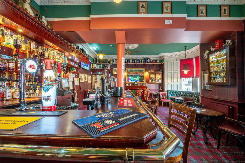 Interior of a cozy pub with a wooden bar counter, beer taps, and seating area with chairs and tables. The walls are decorated with framed pictures and shelves filled with bottles, with green and red color scheme.