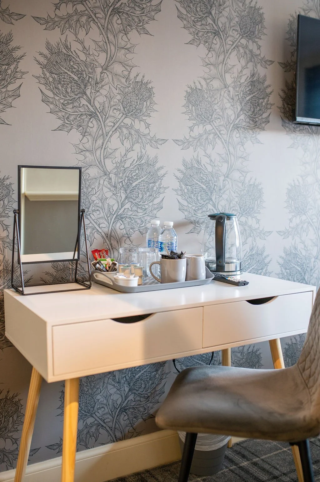 A white desk with a mirror, water bottles, cups, and a kettle in front of a gray floral wallpaper, with part of a TV mounted on the wall to the right and a gray upholstered chair in front of the desk.