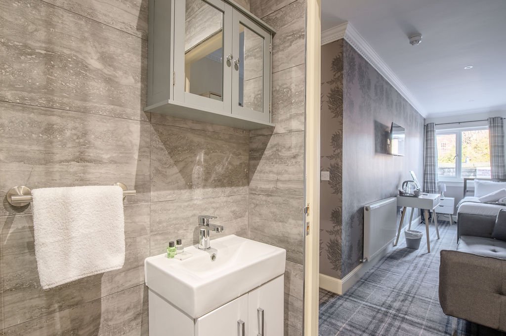 A bathroom with gray tiled walls, a white sink with a faucet, a towel rack with a white towel, and a gray wall-mounted cabinet with a mirror. Adjacent to the bathroom is a living room with patterned wallpaper, a wall-mounted TV, a window with curtain