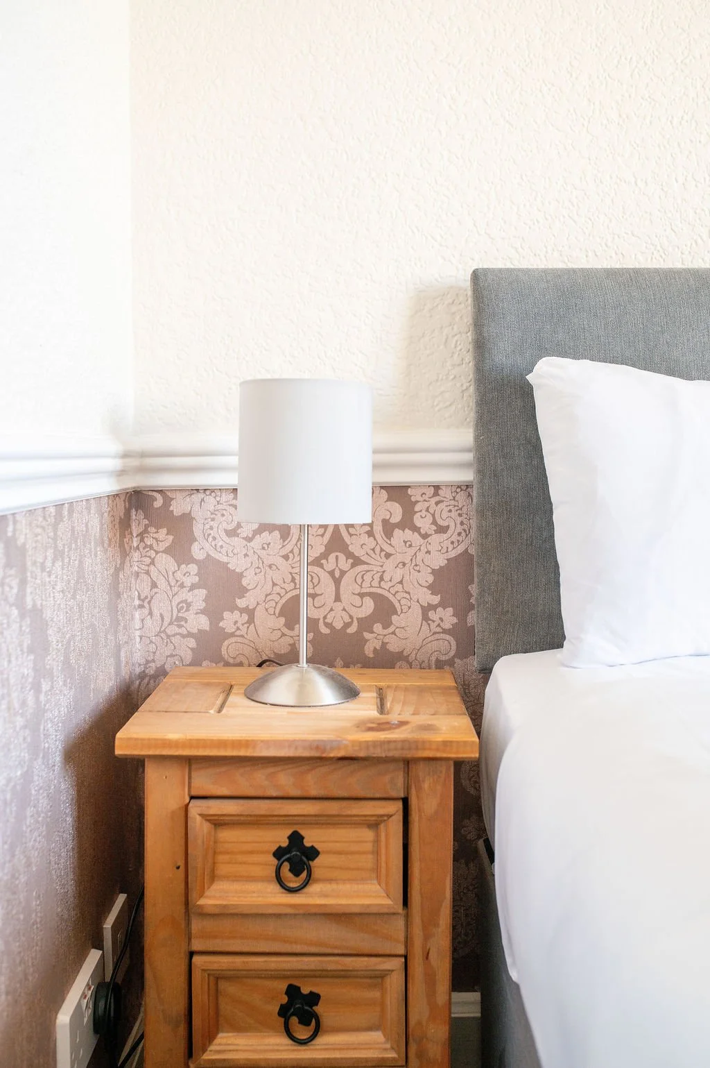 A nightstand with two drawers and black ring pulls, a silver lamp with a white shade, a bed with white bedding and a gray upholstered headboard, and patterned wallpaper behind.