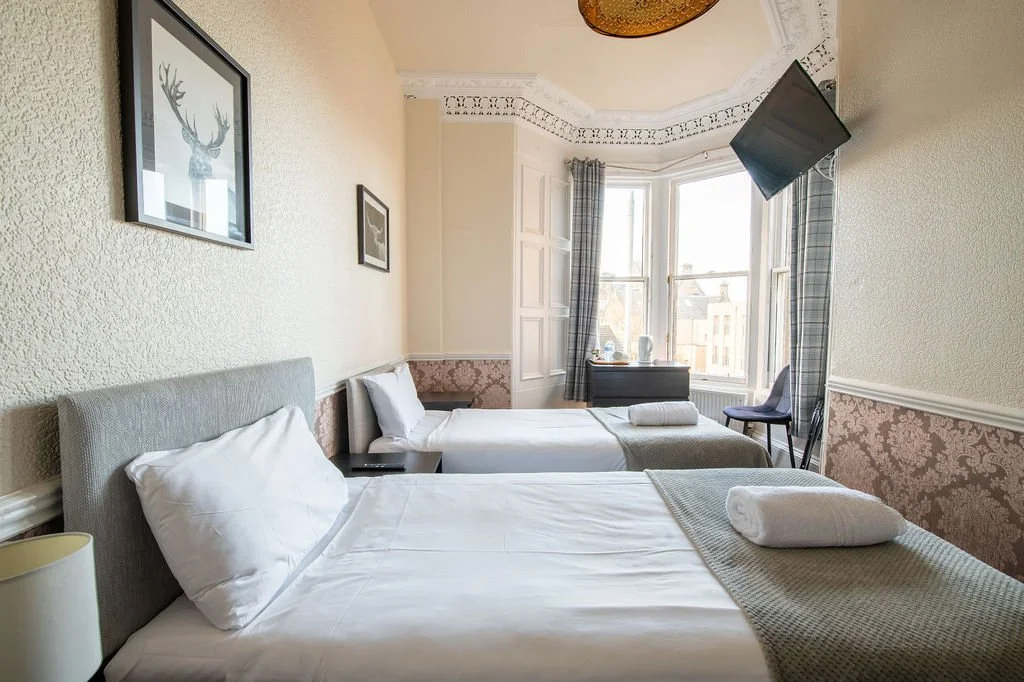 A hotel room with two beds, a window with checkered curtains, a wall-mounted TV, and wall art featuring an elk head.