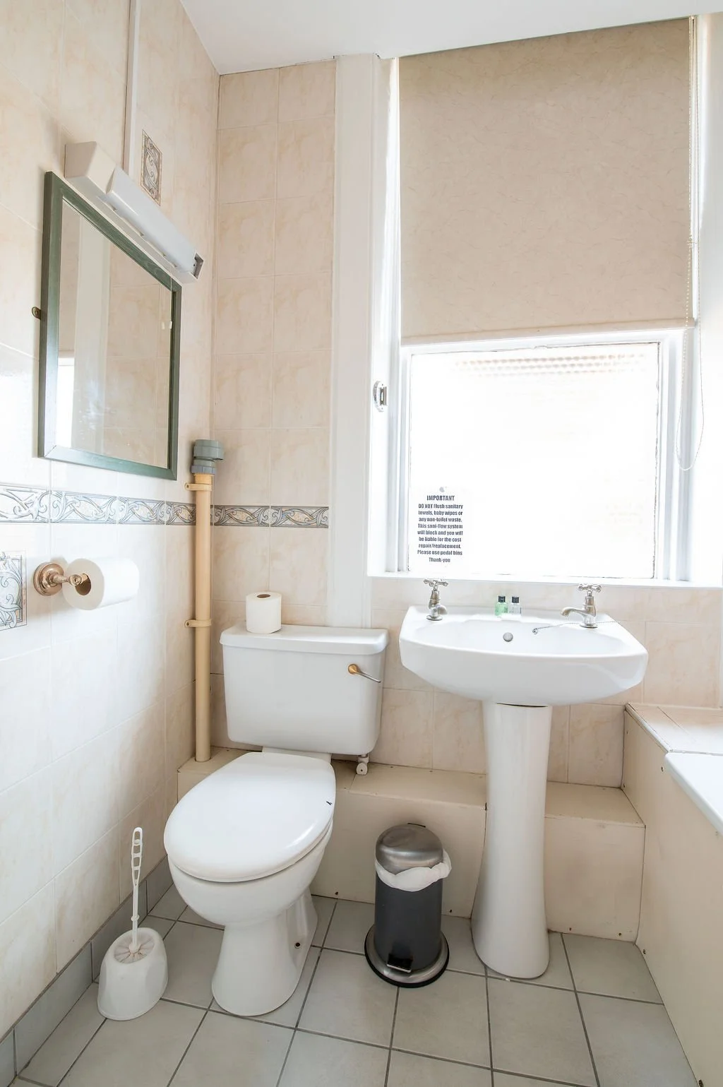 A small bathroom featuring a toilet, a pedestal sink, a rectangular mirror, and a window with a beige roller shade. Toilet paper, a toilet brush, and a trash bin are also visible.