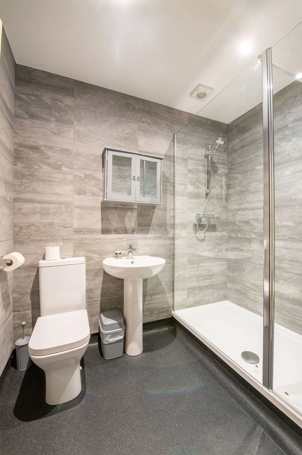 A modern bathroom with a toilet, a pedestal sink, a small wall-mounted cabinet, and a walk-in shower with a glass door. The walls are covered in grey textured tiles, and the floor is dark.