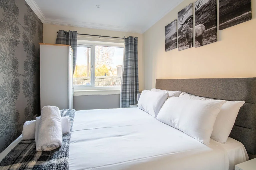A neatly made bed with white sheets and pillows, a rolled towel on a checkered blanket, in a cozy bedroom with patterned wallpaper, a window with plaid curtains, and black and white moose artwork above the bed.