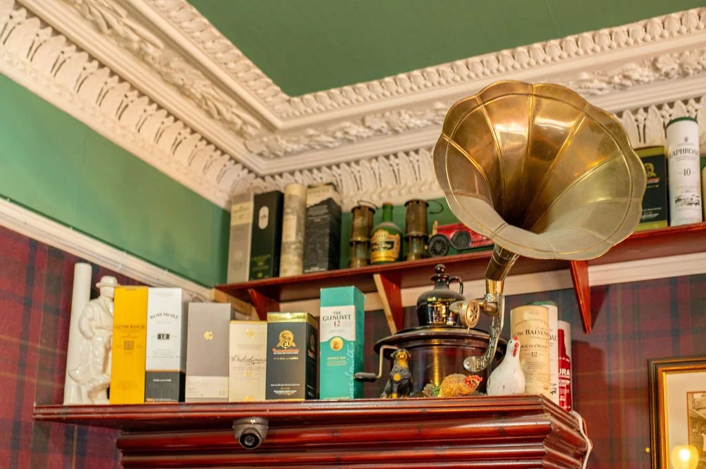 Indoor shelf with a vintage gramophone, various liquor bottles, and figurines, against a green ceiling and patterned wallpaper.