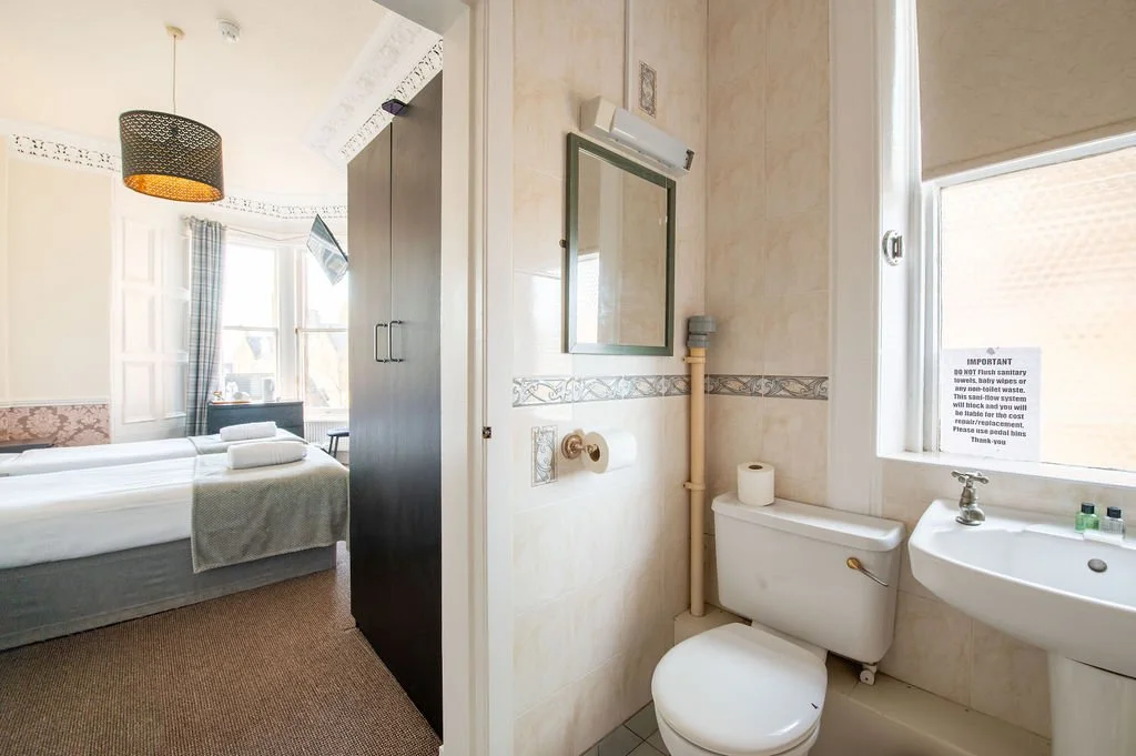 A small bathroom with a toilet, sink, mirror, and a window with a sign, adjacent to a bedroom with a bed and a television.