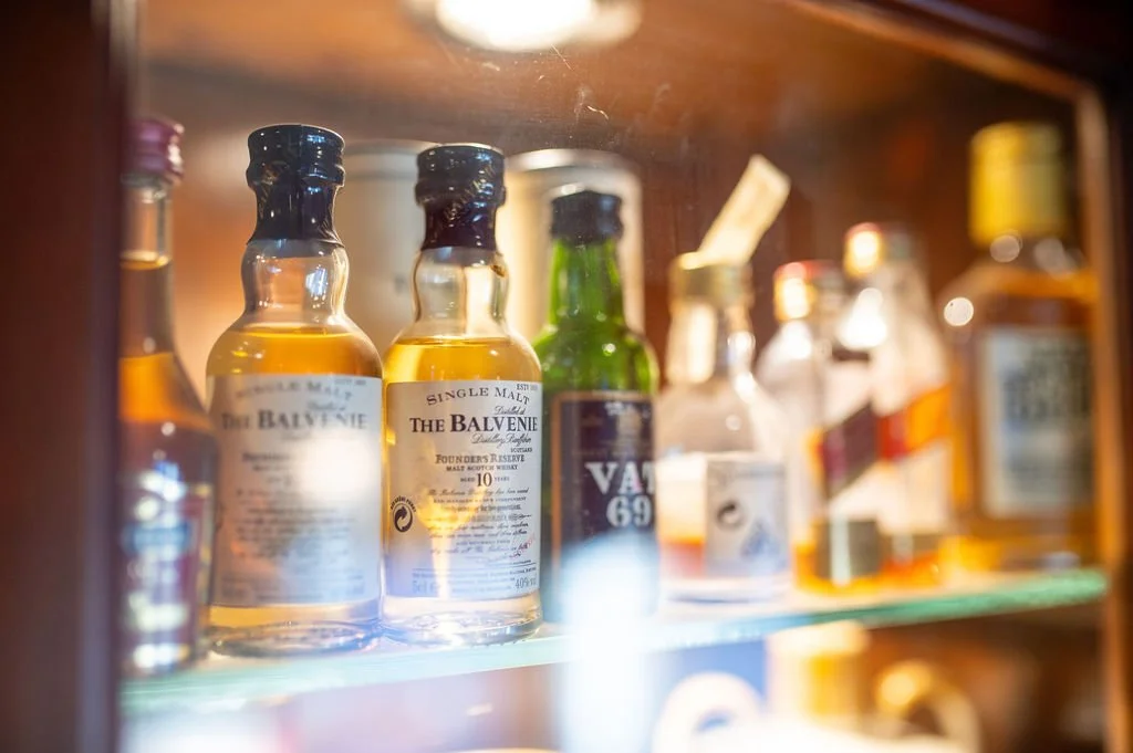 Various bottles of alcohol on a glass shelf inside a wooden cabinet, including two bottles of The Balvenie single malt Scotch whiskey, a green bottle, and other assorted spirits.