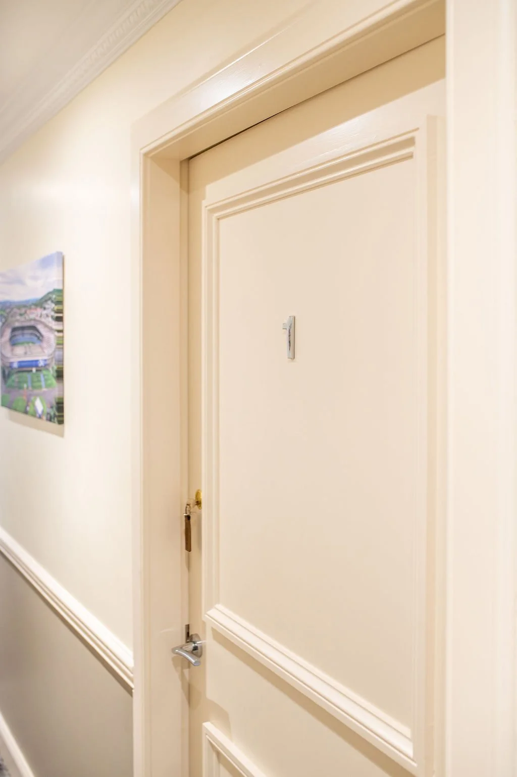 Close-up of an interior door with a peephole, surrounded by cream-colored walls and crown molding, with a picture hanging on the wall to the left.