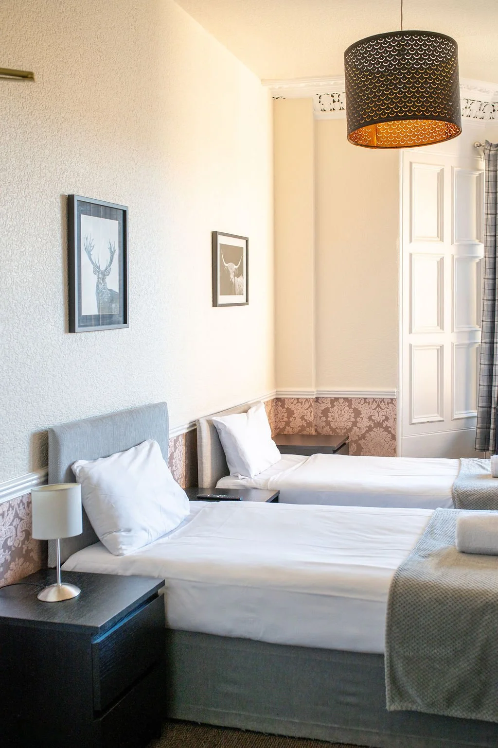 Hotel room with two single beds, white bedding, pillows, bedside tables, framed artwork of deer on the wall, a black ceiling light, curtains, and a patterned wallpaper border.