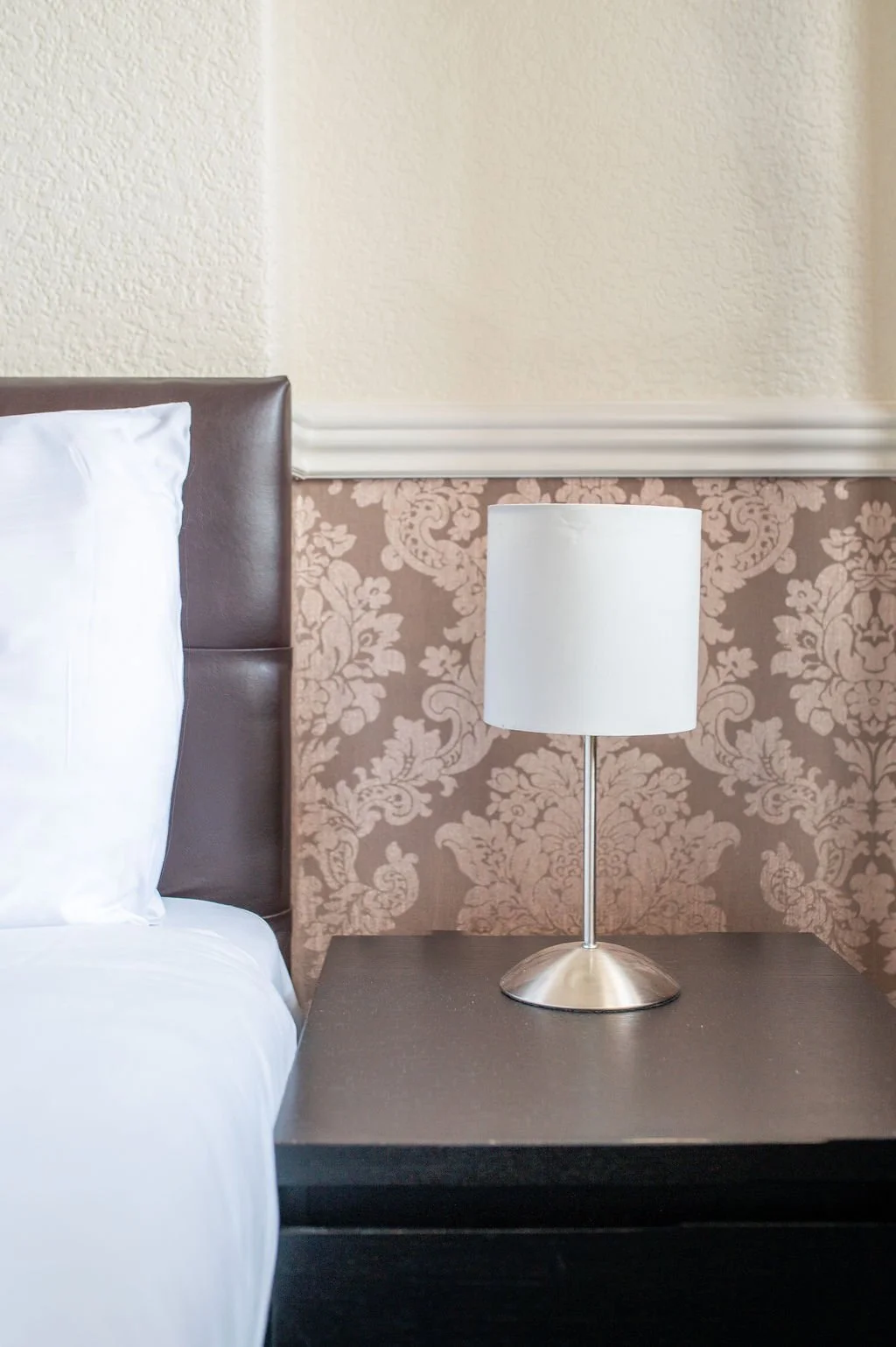 A close-up of a bedroom nightstand with a modern silver table lamp against a patterned wallpaper background.