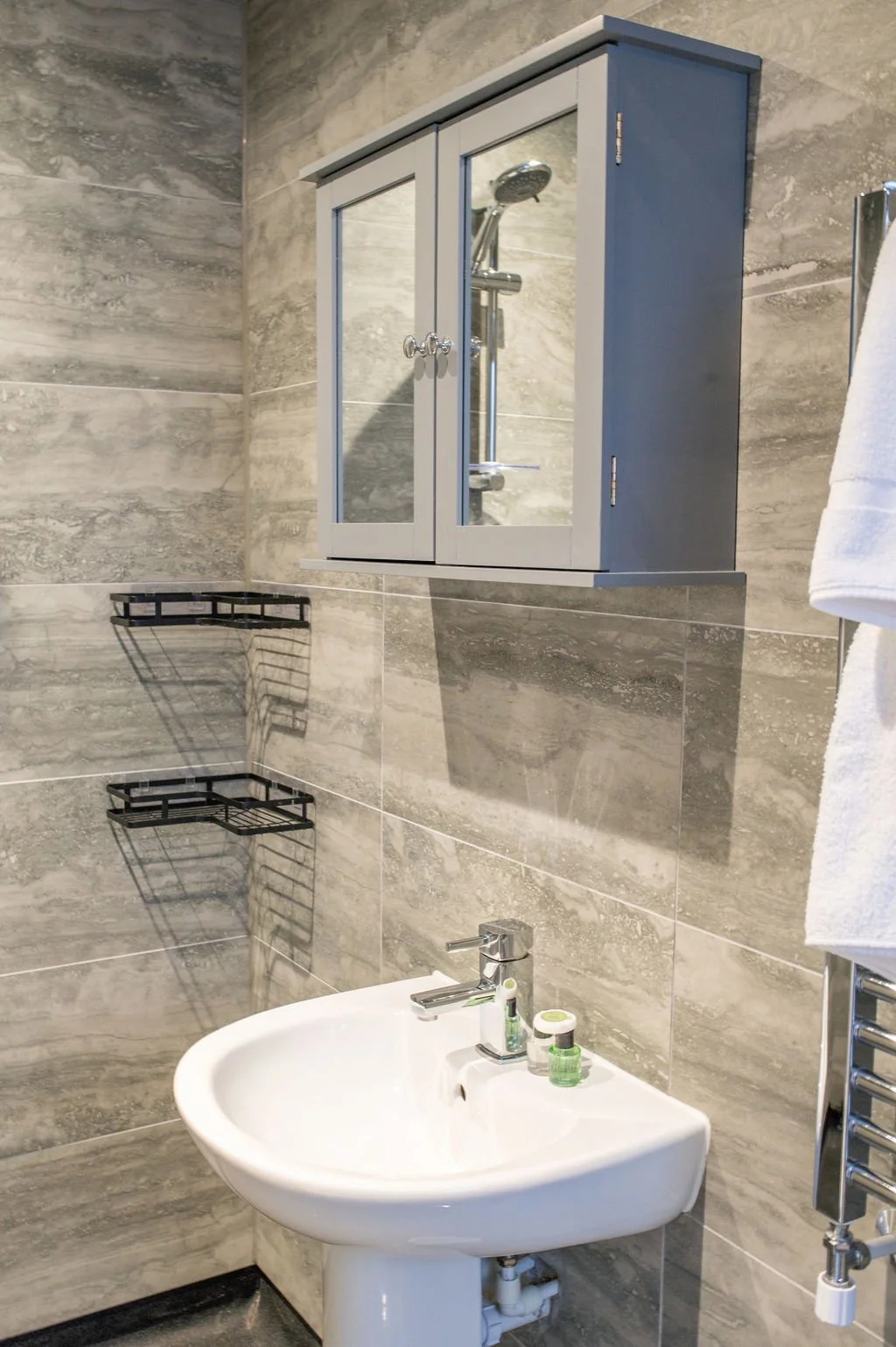 Bathroom with grey tiled walls, a white bidet, a mirror cabinet above the bidet, two black wire shelves, and a white towel hanging on a chrome towel rack.