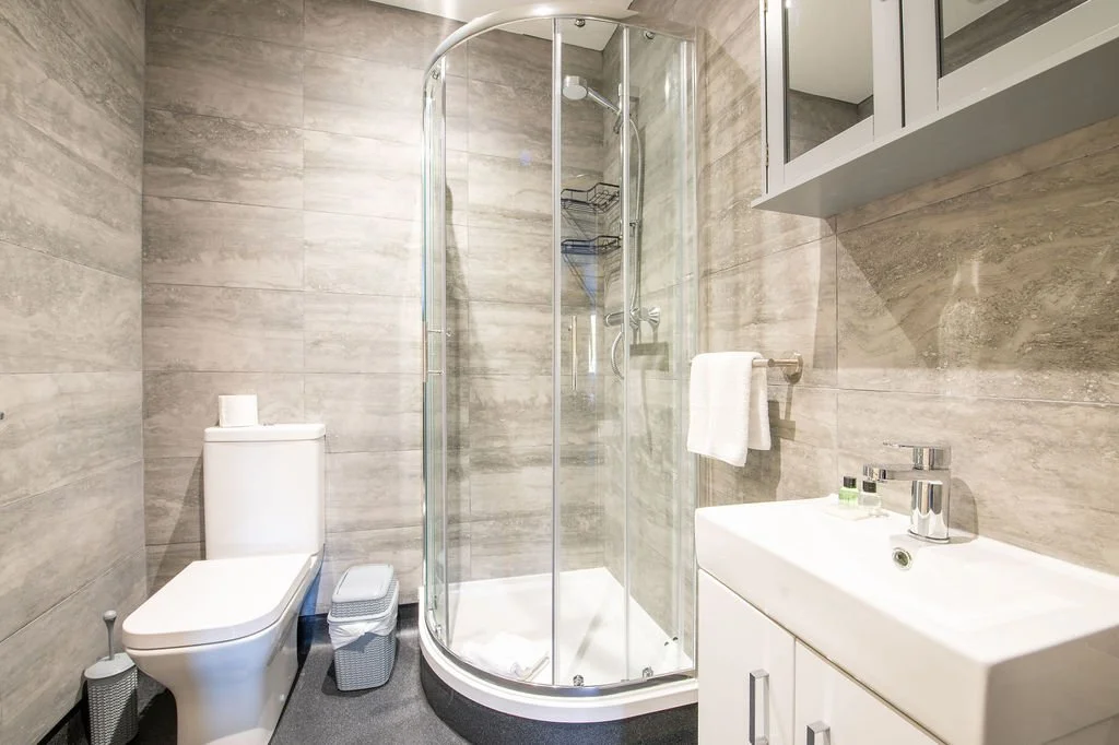 A modern bathroom with a corner shower enclosed by glass, a white toilet, a small white sink with a mirror cabinet above, and neutral-colored tiled walls.