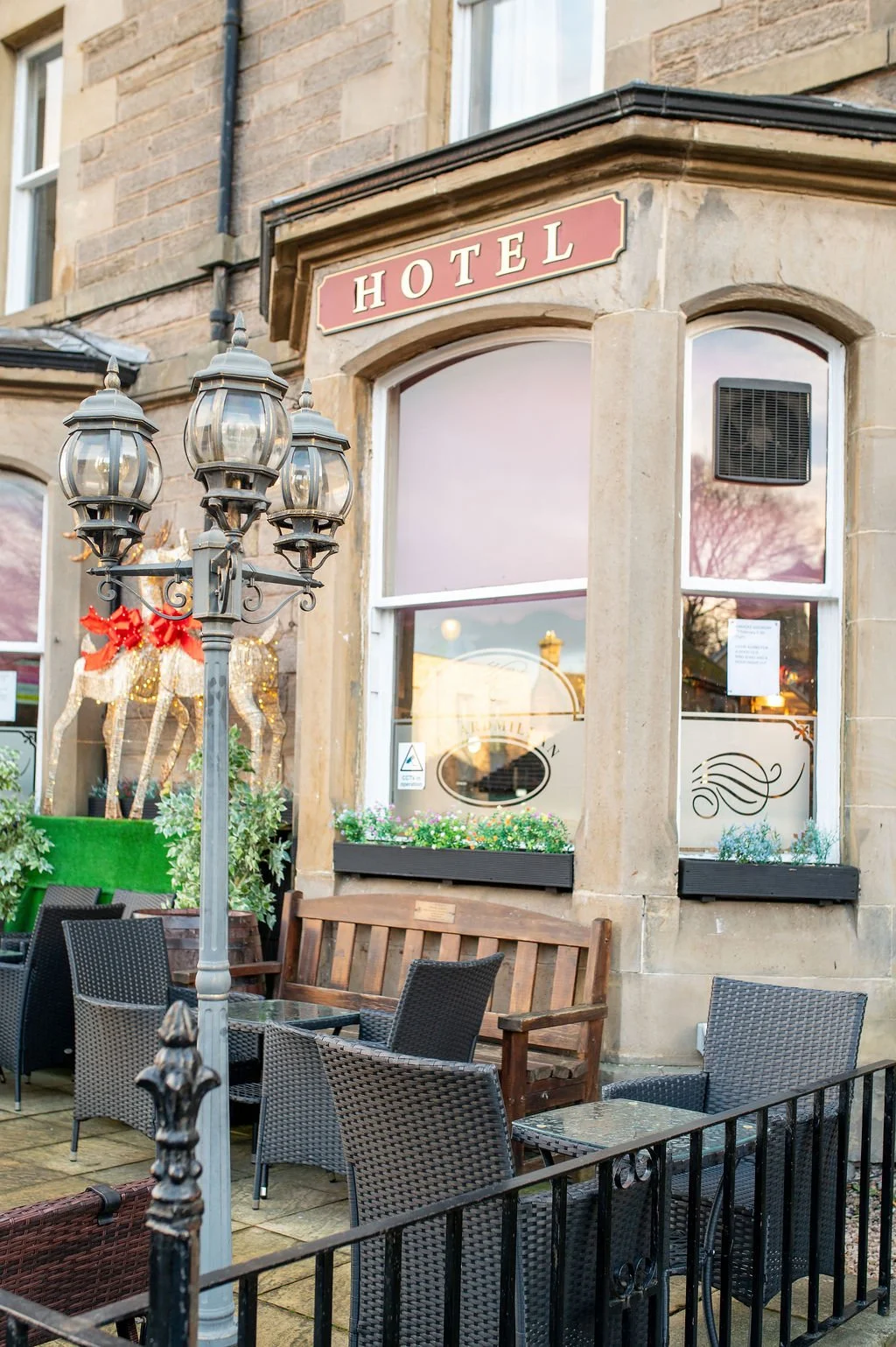 Outside view of a hotel with outdoor seating, a vintage-style street lamp, holiday decorations, and a sign reading 'HOTEL' above the window.