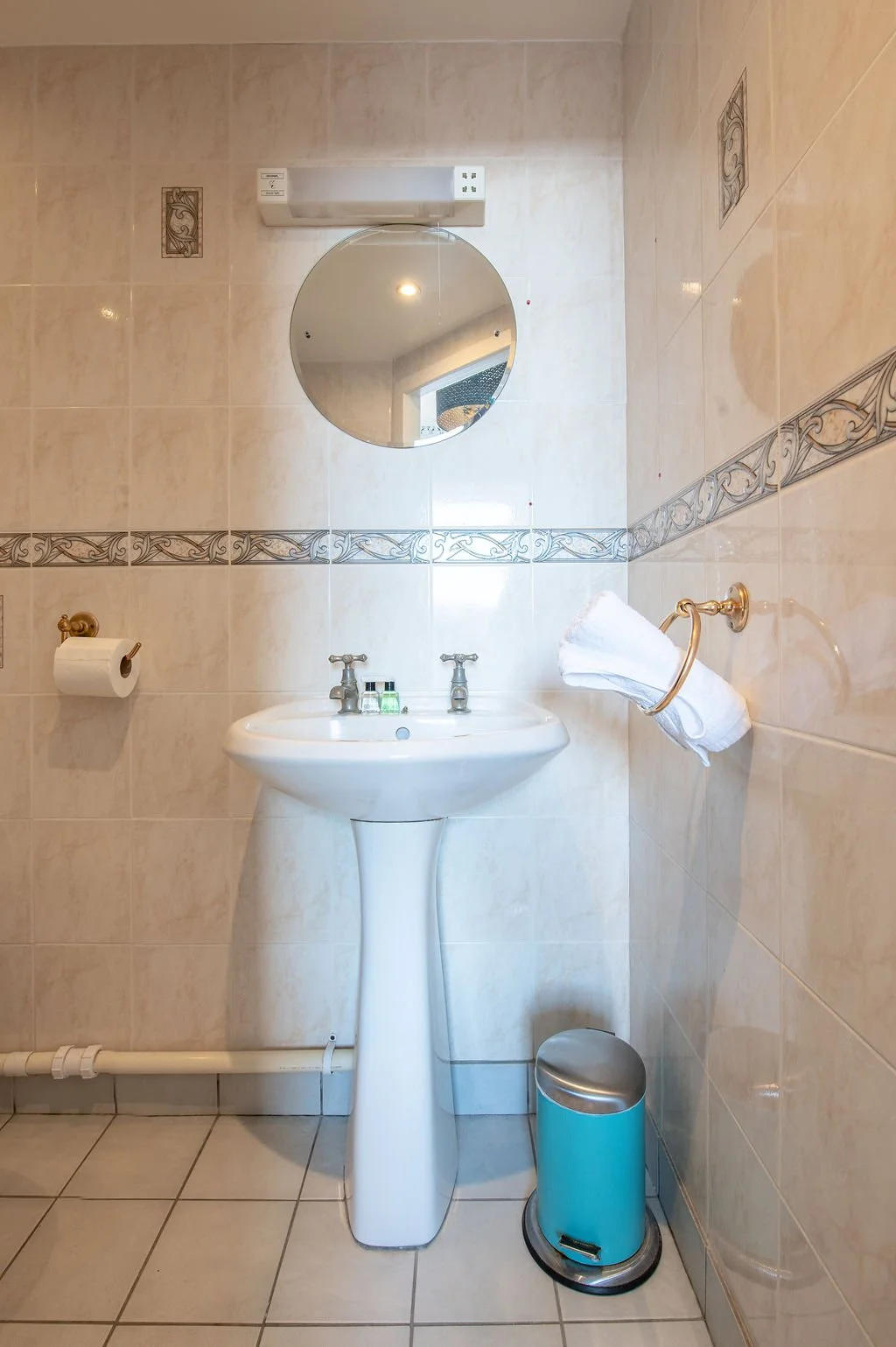 A small bathroom with tiled walls and floor, a pedestal sink with two faucets, a round mirror above the sink, a toilet paper roll on a holder on the left wall, a towel ring with a folded towel on the right wall, a small trash bin on the floor next to
