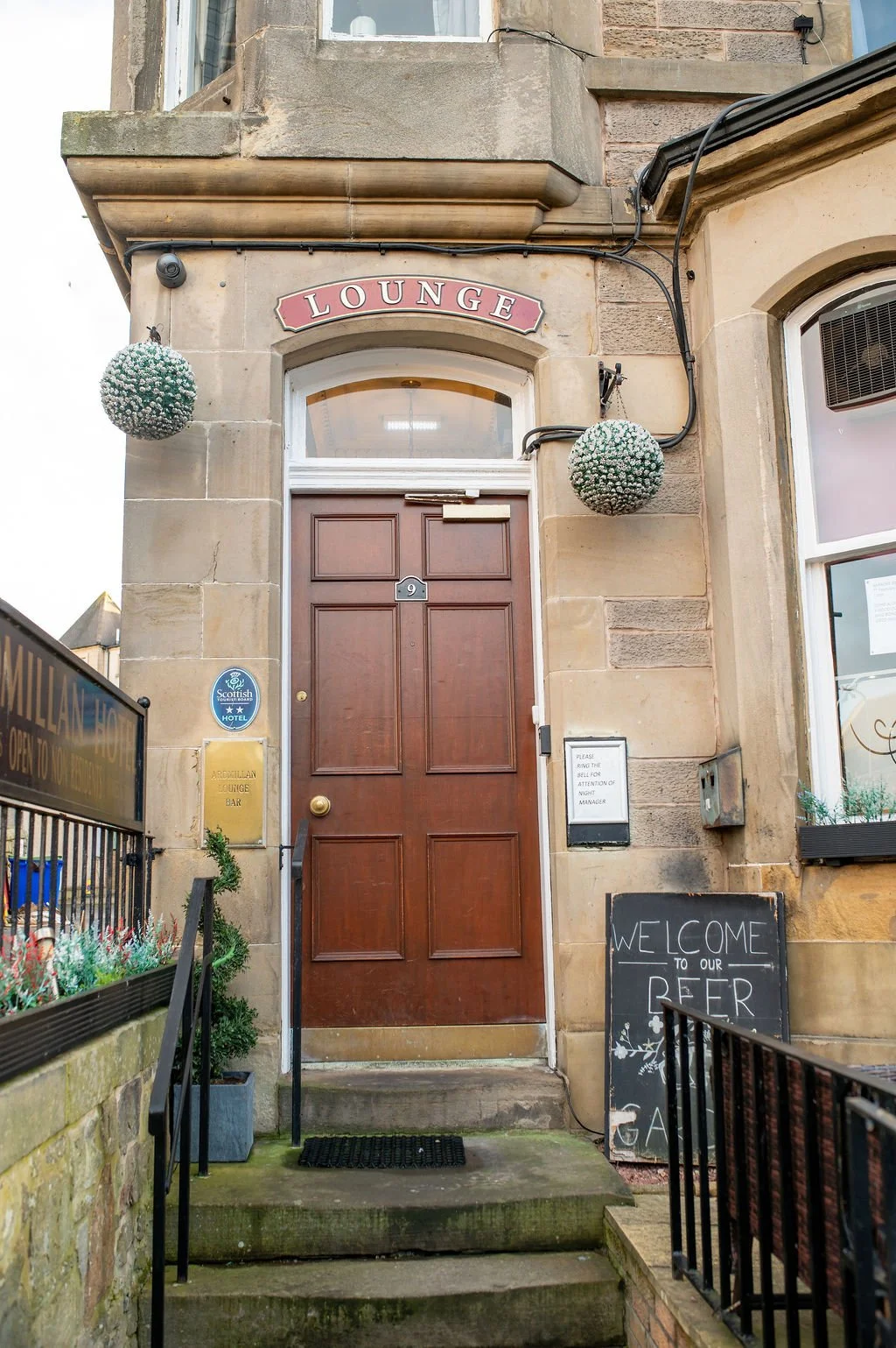Exterior entrance to a lounge or bar with a wooden door, above which is a sign that says 'LOUNGE.' There are hanging decorative balls with white flowers, and a chalkboard sign on the right that says 'WELCOME TO OUR BEER GARDEN.' The area has stone st