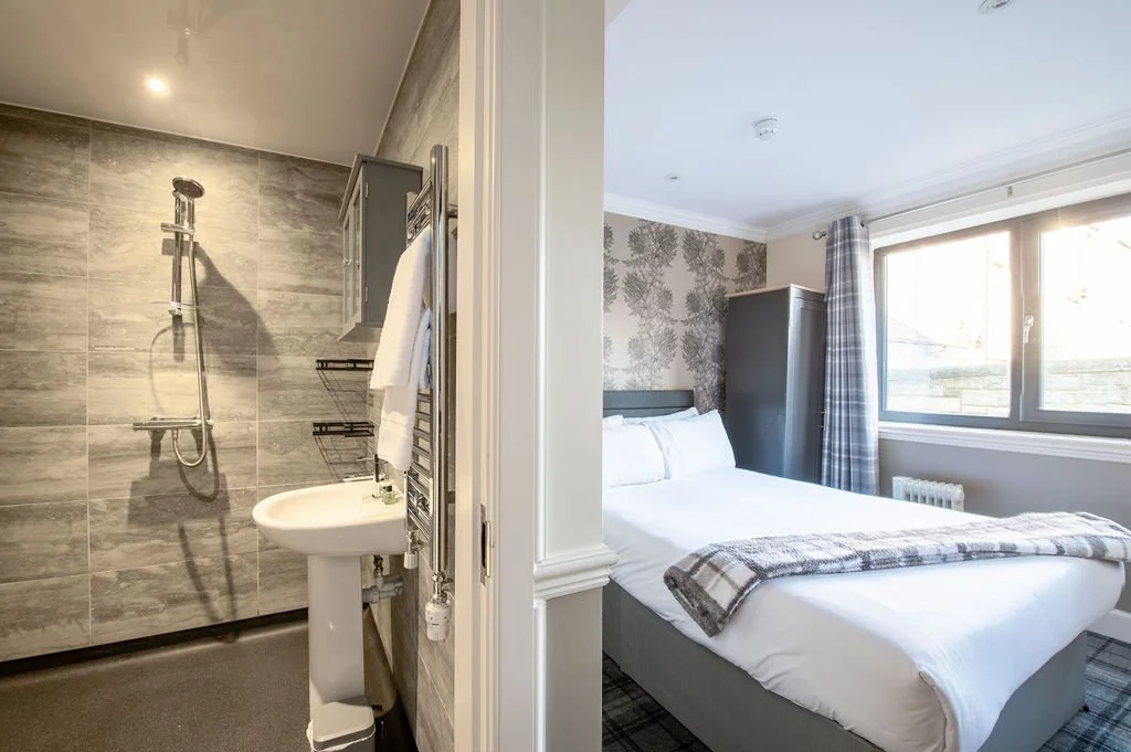 Part of a bathroom with a sink and shower on the left, and a bedroom with a bed, window, and wardrobe on the right.