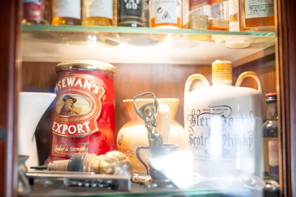 Inside a glass cabinet, various vintage items including a red McEwan's Export tin, a ceramic jug with dark lettering, a small figurine, a white mug, a miniature statue of a woman, and a small glass bottle.