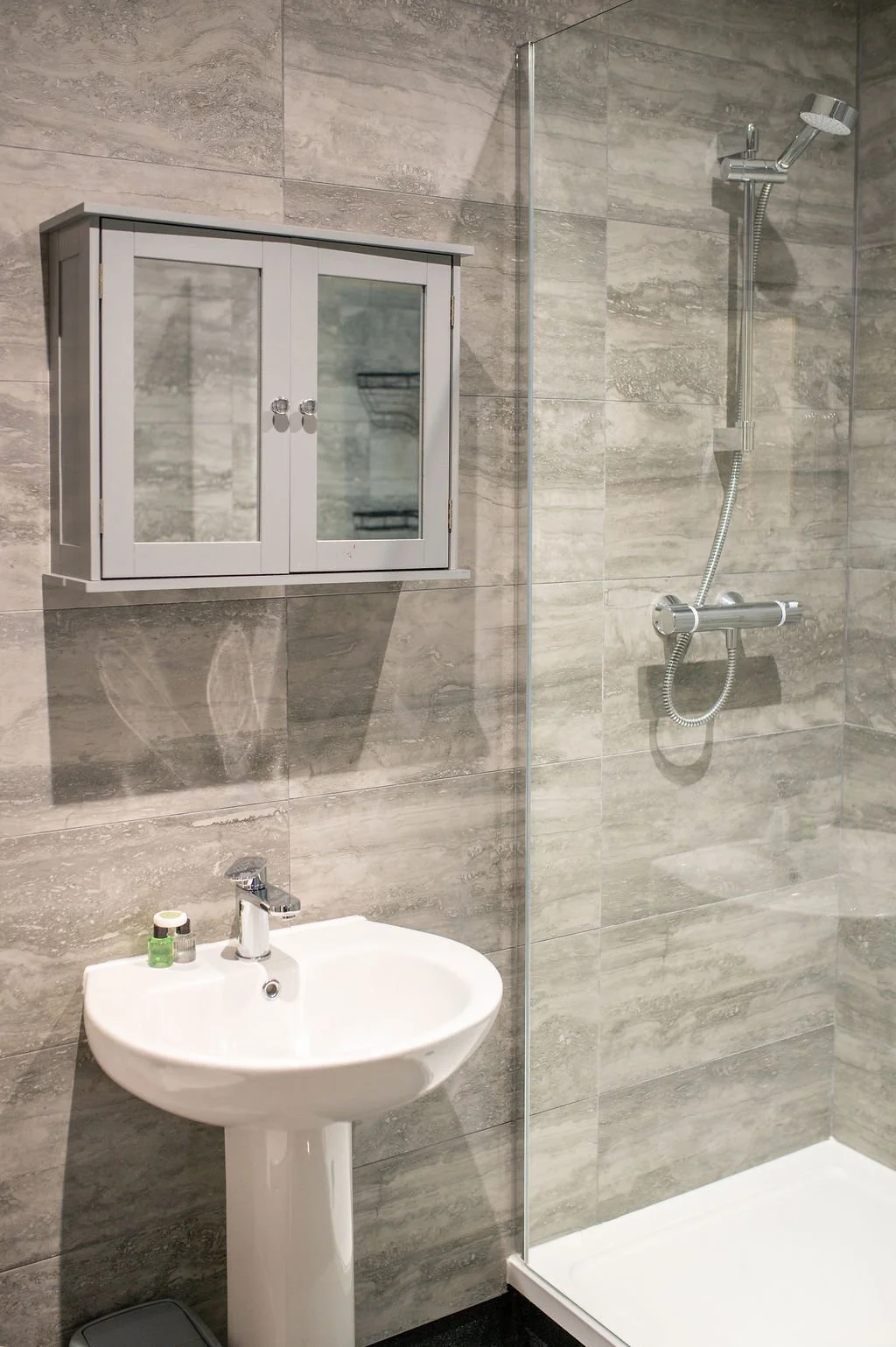 A small bathroom with a gray tiled wall, a white pedestal sink with toiletries, a wall-mounted gray cabinet with mirrored doors, and a shower area with a glass partition and a modern showerhead.