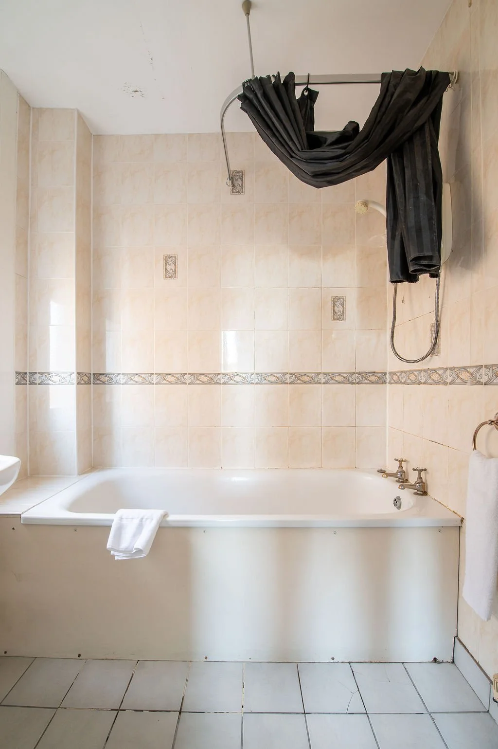 Empty bathroom with beige tiled walls and floor, a white bathtub with a towel draped over it, and black curtains hanging from a curved shower rod.