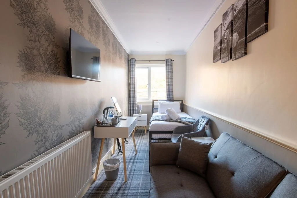 Hotel room with a wall-mounted TV, a white desk with a kettle, cups, and water bottles, a single bed with white linens, a gray armchair, a dark gray sofa with cushions, a window with plaid curtains, and wall art.