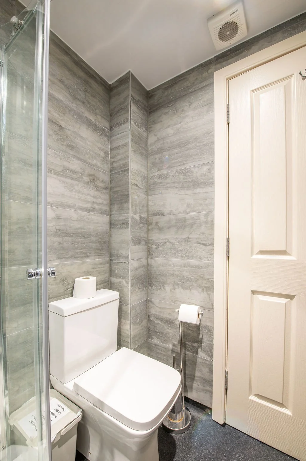 A small bathroom with gray tiled walls, a white toilet, a toilet paper roll on a stand, a small trash bin, and a partially visible shower enclosure with a glass door. There is a white door and a ceiling vent.
