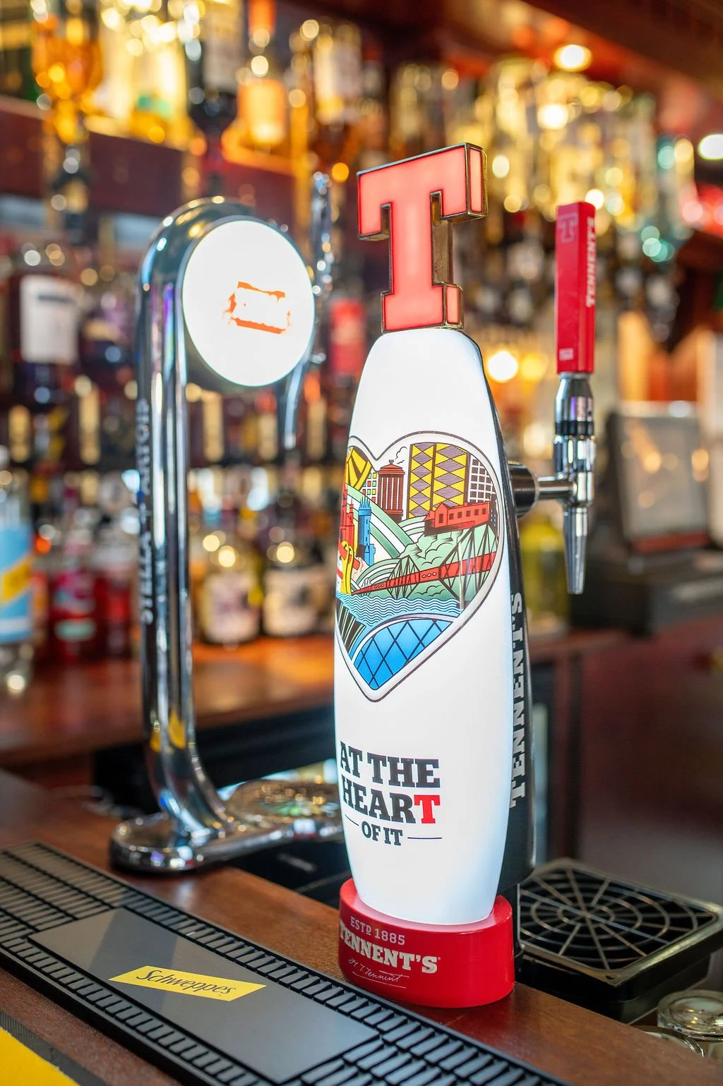 Beer tap handle with a colorful design and the phrase 'At the Heart of It' in a pub or bar setting.
