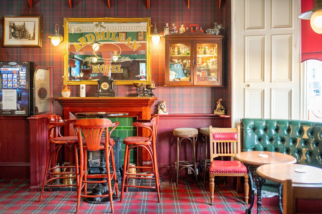 Cozy pub interior with red plaid carpet, green leather Chesterfield sofa, wooden chairs, and a bar area with stools, a mirror, and vintage decor.