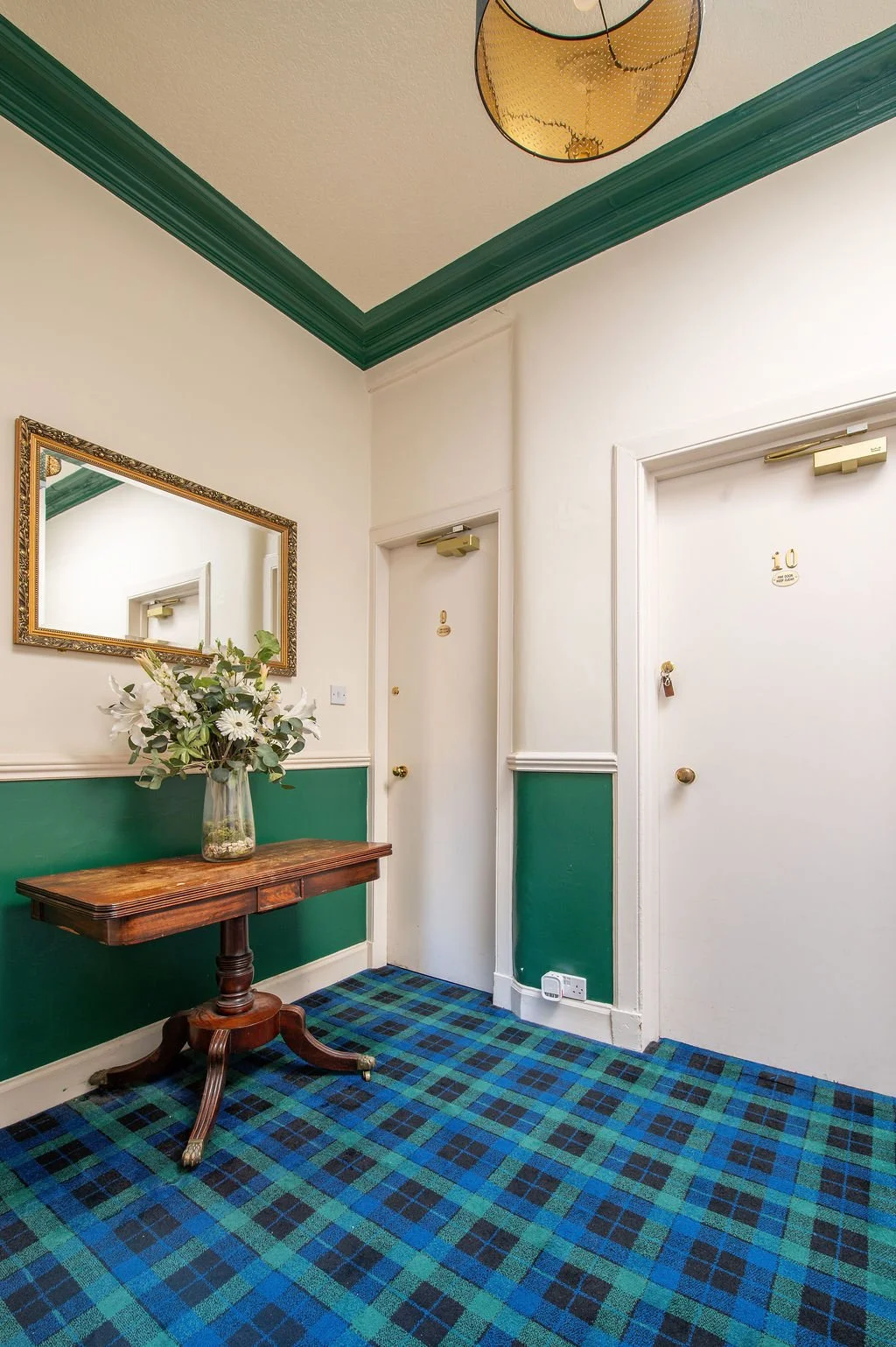 A hallway corner with a wooden table holding a clear glass vase with white flowers and green foliage, a large framed mirror on the wall, white door with gold hardware, and a blue and green tartan carpet.