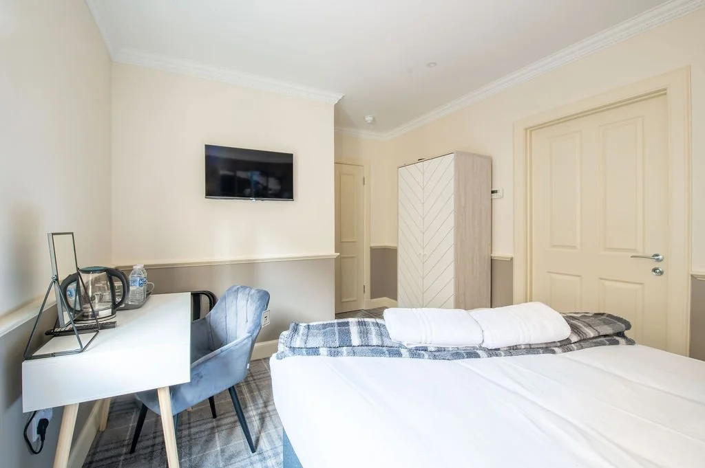 A hotel room with a bed, a desk with a mirror and water, a chair, a wardrobe, and a wall-mounted television.