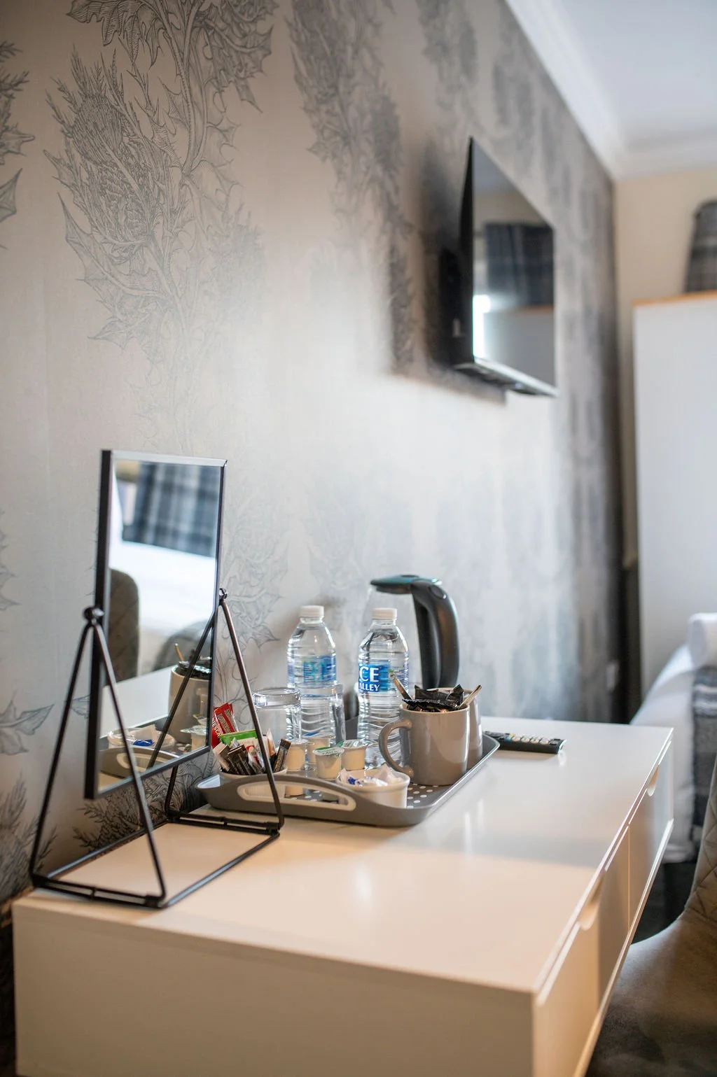 Hotel room workspace with a white desk holding water bottles, cups, and snacks, a small mirror, and a kettle, with a television mounted on a patterned wallpapered wall.