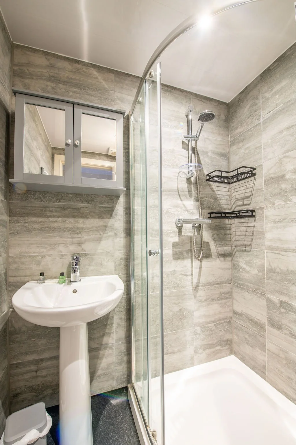 Small bathroom with a shower, pedestal sink, gray storage cabinet with mirror, and gray tiled walls.