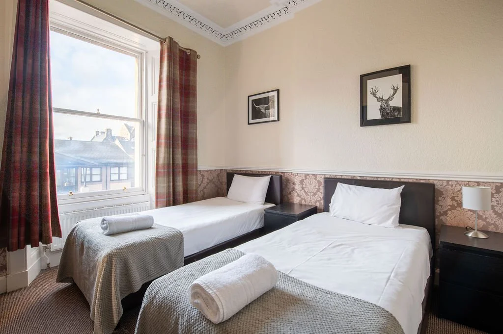 A bedroom with two single beds, each with white sheets and towels, dark wood nightstands, and framed pictures of a deer and a moose on the wall. A large window with plaid curtains shows rooftops outside.