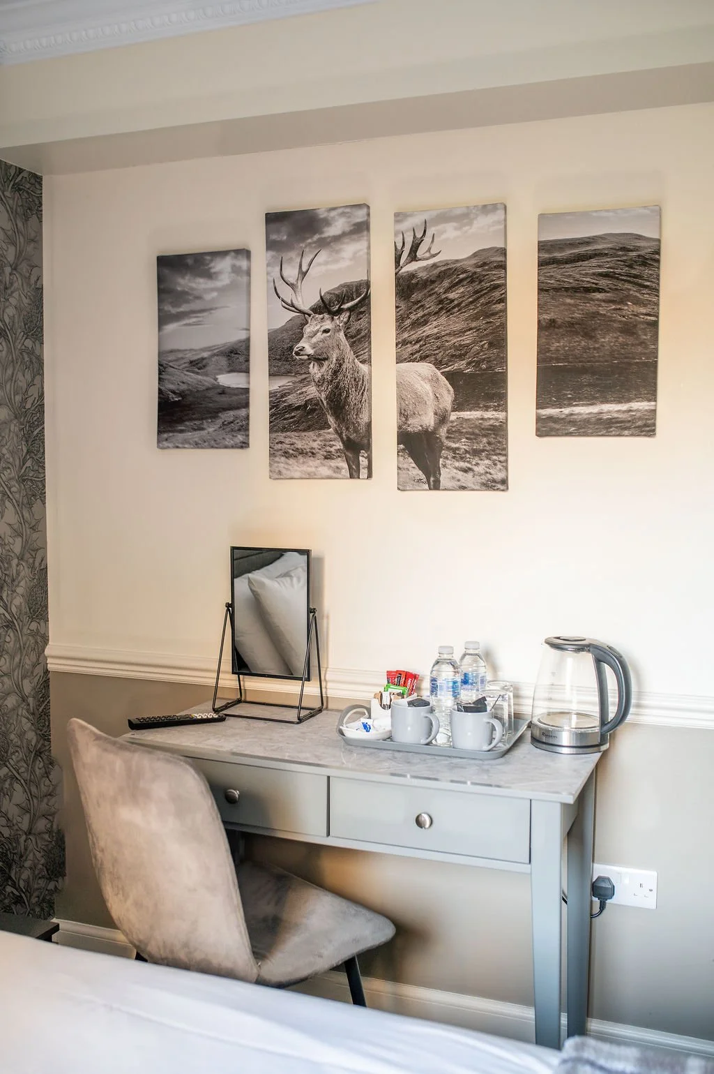 A hotel room corner with a desk holding a mirror, water bottles, cups, and a kettle, with a beige chair in front and four black and white landscape prints of a stag and mountains on the wall.