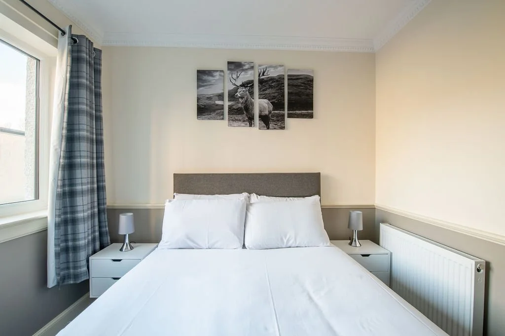 A neatly made bed with white sheets and pillows, two nightstands with small lamps, a window with plaid curtains, and a multi-panel framed black and white picture of a deer in a landscape above the bed.