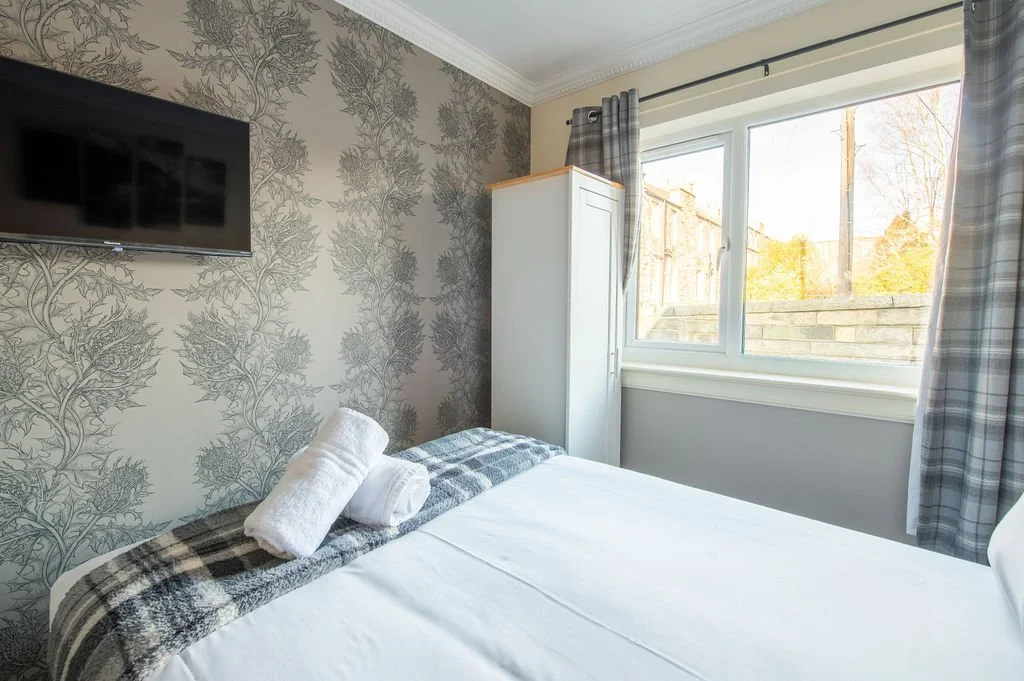 A cozy bedroom with a bed, a white wardrobe, a mounted flat-screen TV, and a window with gray plaid curtains showing an outdoor view of trees and a brick wall.