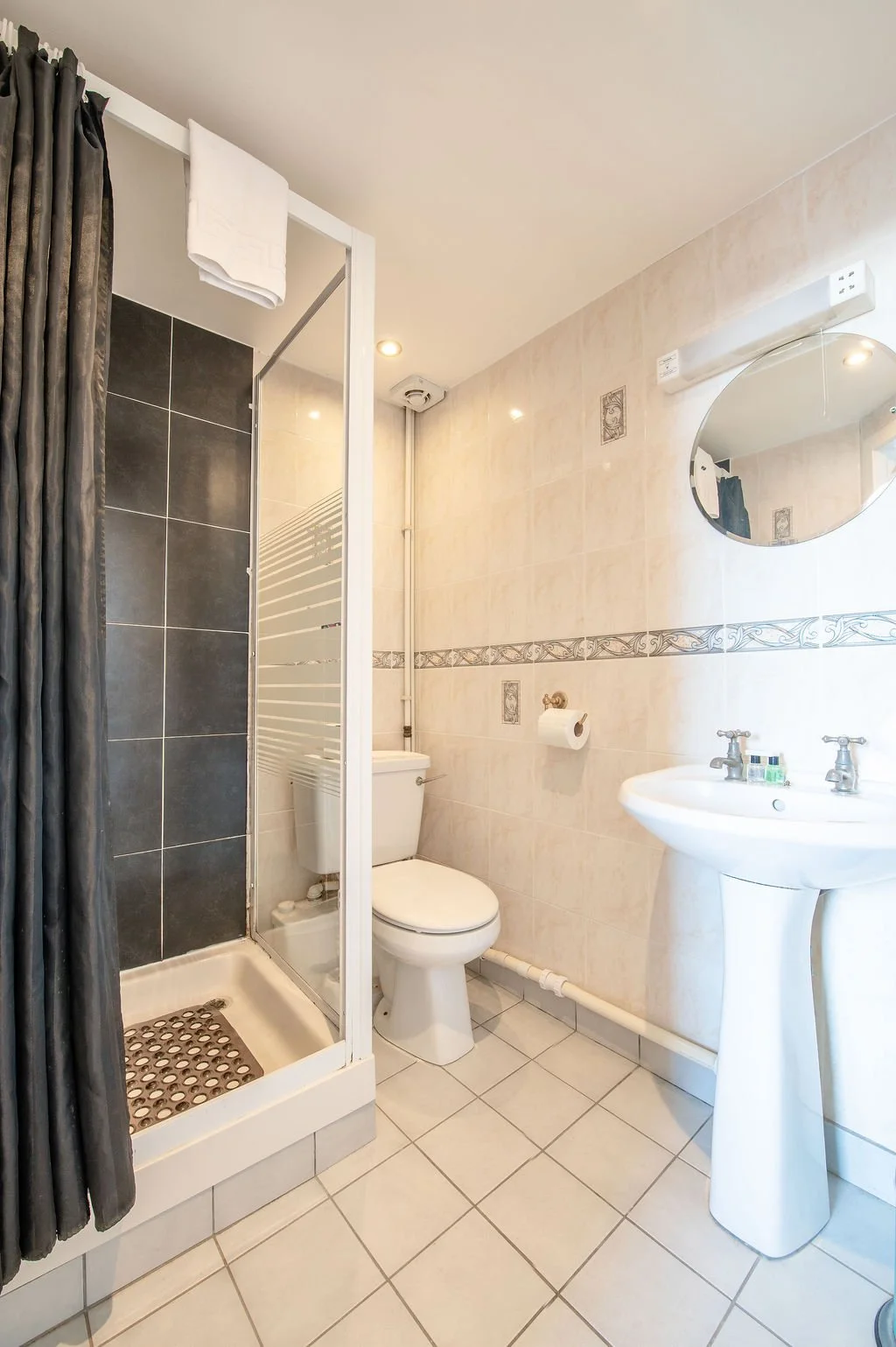 A small bathroom with a shower stall, toilet, and pedestal sink. The shower has dark tiles and a curtain, while the walls are light-colored with decorative border tiles. A round mirror hangs above the sink, and there is a toilet paper holder on the w